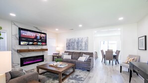 A modern living room with a flat-screen TV displaying a movie, a fireplace, a sofa, a coffee table, and a dining area with chairs and a table. - Modern clean relaxing gateway! Close to all LA fun! (Monterey Park)