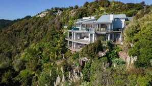 A large, modern house with multiple levels, surrounded by dense forest and hills.