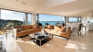 A spacious living area with two leather sofas, a glass coffee table, and a dining area with a white table and chairs. Large windows offer a view of the sea and hills.