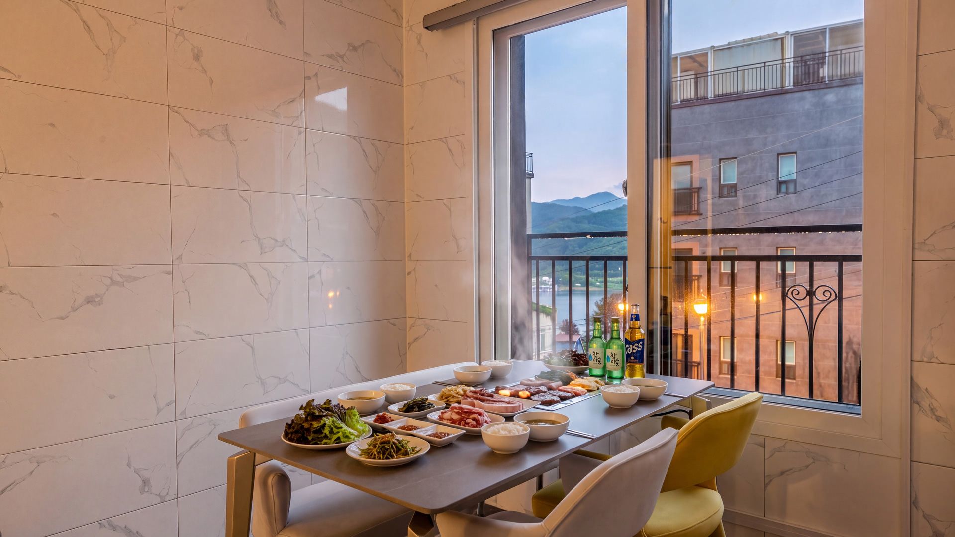 Un comedor con una mesa dispuesta para una comida, una ventana con vistas a edificios y montañas, y un balcón con barandilla.
