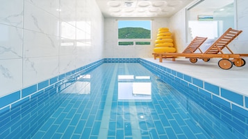 Una piscina interior con baldosas azules brillantes, vistas a la vegetación por las ventanas y sillones de madera con ruedas.