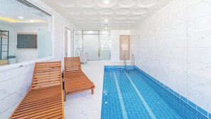 Une piscine intérieure moderne, dotée de chaises longues en bois et d’un espace douche clos par des parois de verre.