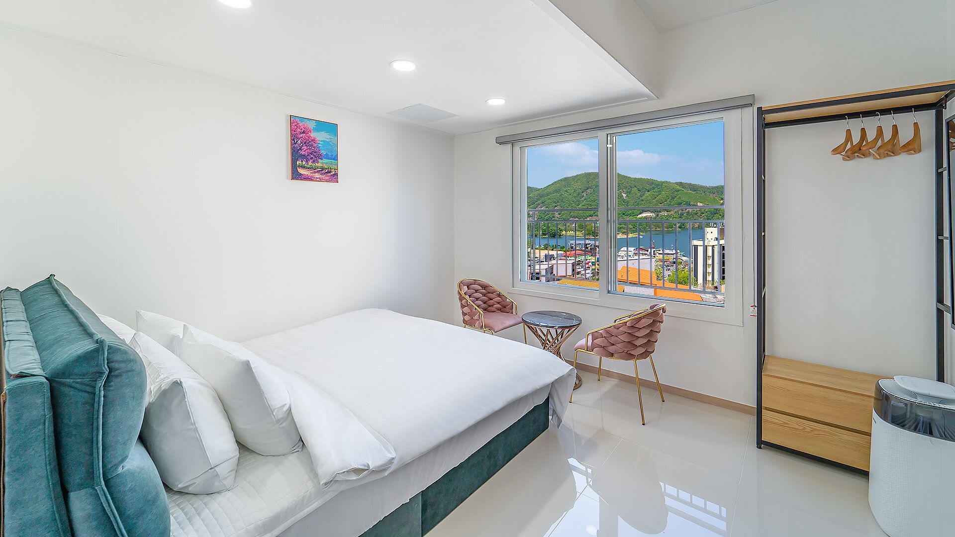 Un dormitorio moderno con una cama, dos sillas, una mesita y un amplio ventanal con vistas a un paisaje urbano.