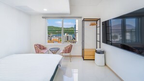 A modern hotel room with a bed, two chairs, a small table, a TV, and a balcony with a view of the city and hills.
