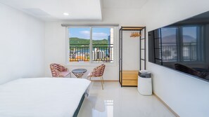 A modern hotel room with a bed, two chairs, a small table, a TV, and a balcony with a view of the city and hills. - Gapyeong Boreum-dal Poolvilla (Gapyeong)