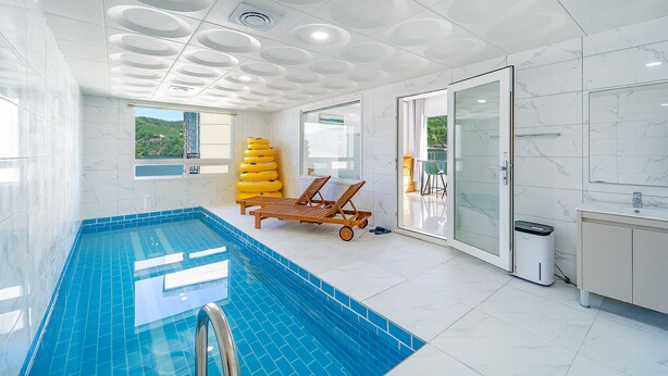 Een modern binnenzwembad met een helderblauw watergedeelte, een houten lounge stoel en een glazen deur naar een balkon.