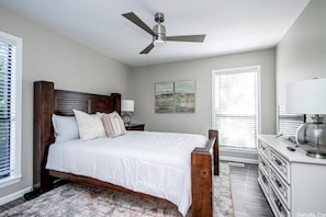 Quarto com cama de madeira, ventilador de teto, escrivaninha com abajur e janela com persianas.