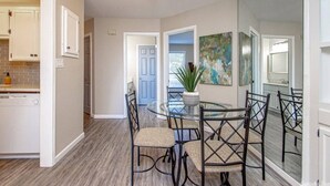 Un comedor con mesa y sillas de vidrio, una planta en maceta y vistas de la cocina y la sala de estar.