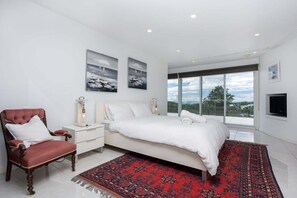Une chambre moderne avec un grand lit, un fauteuil rouge et une vue sur l’extérieur grâce à de grandes fenêtres.