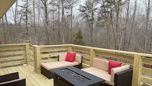 Une terrasse en bois, meublée de mobilier en osier, et dotée d’un foyer intégré.