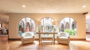 Amplia habitación con piso de madera, dos sillones blancos, una mesita y ventanas arqueadas grandes que ofrecen vistas a plantas en macetas y un edificio con arcos.