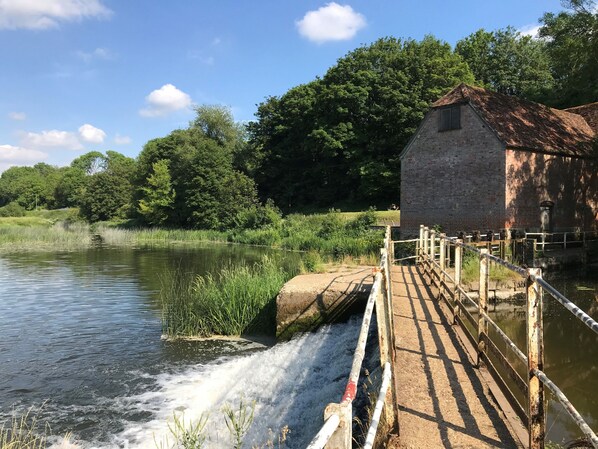 A rustic water mill with a wooden bridge over a flowing stream, surrounded by lush greenery and a clear blue sky.