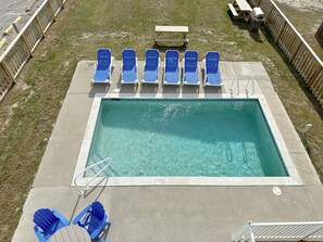 Une piscine rectangulaire à l’eau claire, entourée d’une terrasse en béton. On y trouve six chaises longues bleues disposées en deux rangées de trois, et une table de pique-nique en bois au centre.