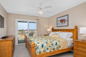 A bedroom with a bed, a desk, a chair, a ceiling fan, a nightstand, a lamp, a window, and a door leading to a balcony. - Turning Tide: Semi-Oceanfront, private pool & hot tub, rec room with pool table! (Nags Head)