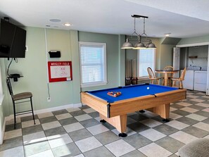 A pool table in a room with a checkered floor, a television mounted on the wall, and a window with blinds.