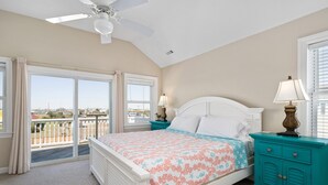 Une chambre à coucher avec un lit, deux tables de chevet, un ventilateur de plafond et une vue dégagée grâce à de grandes fenêtres.