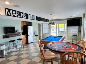 A living room with a pool table, bar stools, a television, and a kitchen area. - Turning Tide: Semi-Oceanfront, private pool & hot tub, rec room with pool table! (Nags Head)