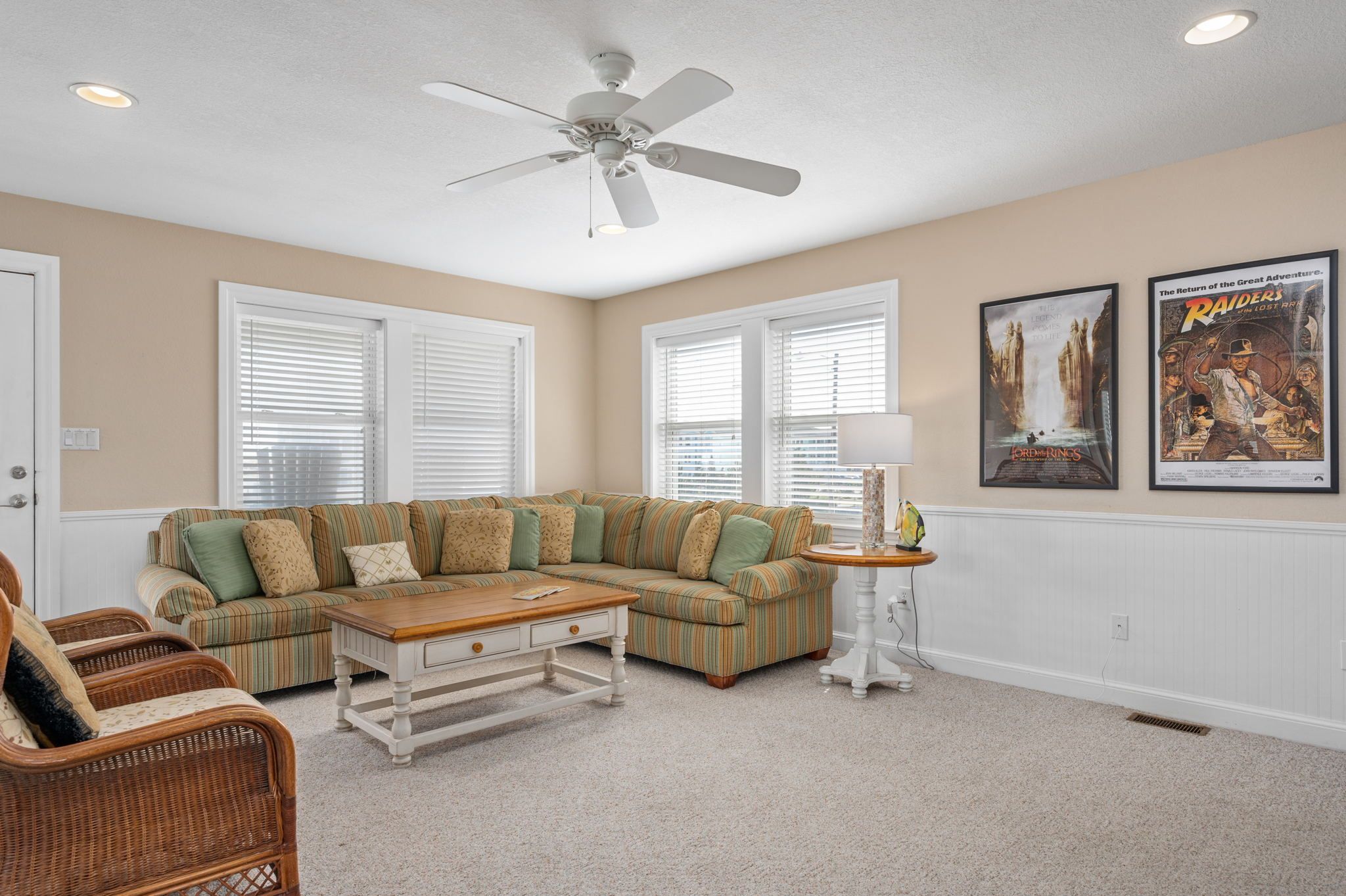 A living room with a sectional sofa, a coffee table, wicker chairs, a ceiling fan, and two framed movie posters.