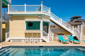 A pool area with a two-story building, a staircase, and a deck with lounge chairs. - Turtle's Nest: Oceanfront, pool, hot tub, theater & rec rooms and elevator! (Kill Devil Hills)