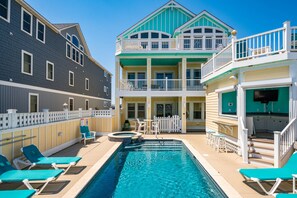 A pool area with lounge chairs, a hot tub, and a two-story building with balconies. - Turtle's Nest: Oceanfront, pool, hot tub, theater & rec rooms and elevator! (Kill Devil Hills)