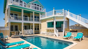 A pool area with a two-story building, a staircase, and lounge chairs. - Turtle's Nest: Oceanfront, pool, hot tub, theater & rec rooms and elevator! (Kill Devil Hills)