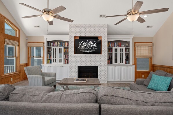 A spacious living room with a fireplace, two ceiling fans, a large flat-screen TV mounted on a brick wall, and a bookshelf filled with books.