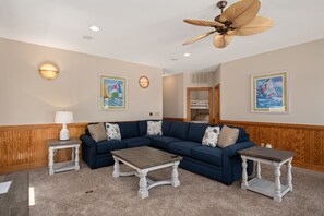 A living room with a sectional sofa, two side tables, a ceiling fan, and artwork on the walls. - Turtle's Nest: Oceanfront, pool, hot tub, theater & rec rooms and elevator! (Kill Devil Hills)