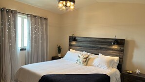 A neatly made bed with white linens, a dark wooden headboard, and a black throw blanket. There is a small bedside table with a lamp and a decorative item. - Luxury Stay w/Coffee Bar & Movie Room, 2 King Suites, Firepit — Black Hills View (Rapid City)