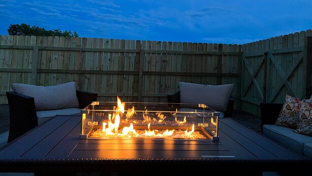 A rectangular fire pit with flames, surrounded by a wooden fence, and two cushioned chairs with pillows. - Luxury Stay w/Coffee Bar & Movie Room, 2 King Suites, Firepit — Black Hills View (Rapid City)