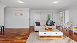 Un salon moderne avec un plancher en bois, un canapé blanc agrémenté de coussins à motifs, une table basse sur laquelle se trouve un saladier rempli de fruits, et un tableau encadré accroché au mur.