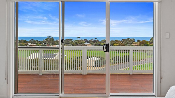 Une pièce dotée d’une grande porte-fenêtre coulissante donnant sur un balcon avec une terrasse en bois et une vue sur l’océan et la végétation.