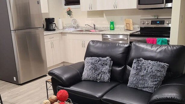Ett modernt kök med vita skåp, ett kylskåp i rostfritt stål, en mikrovågsugn och en ugn. Där finns en svart lädersoffa med grå och blå mönstrade kuddar, ett litet träbord med en korg full av röda och beige bollar samt en blå matta.