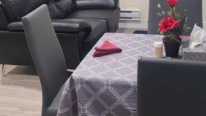 Ett rum med ett matbord, stolar, en svart lädersoffa, en klocka på väggen och två fönster med persienner.