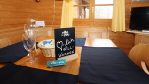 Interior de una cabaña de madera con una mesa de comedor preparada para dos, una cesta, un cartel y un televisor.