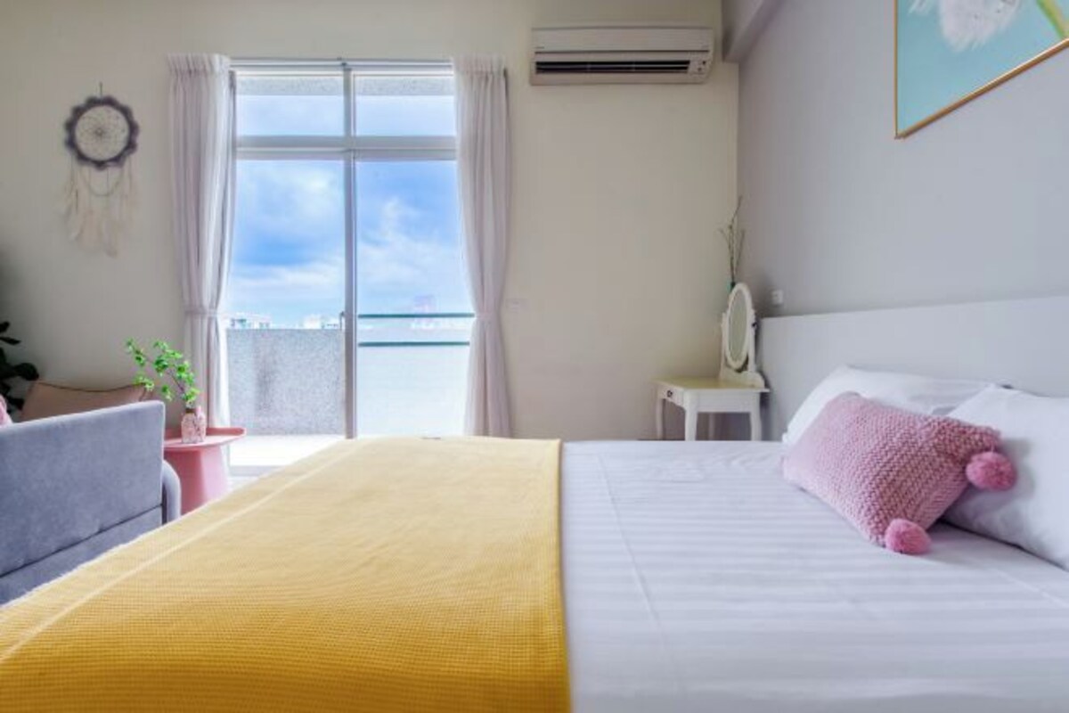 A hotel room with a bed, a bedside table, a chair, a window with curtains, and a view of the sky.