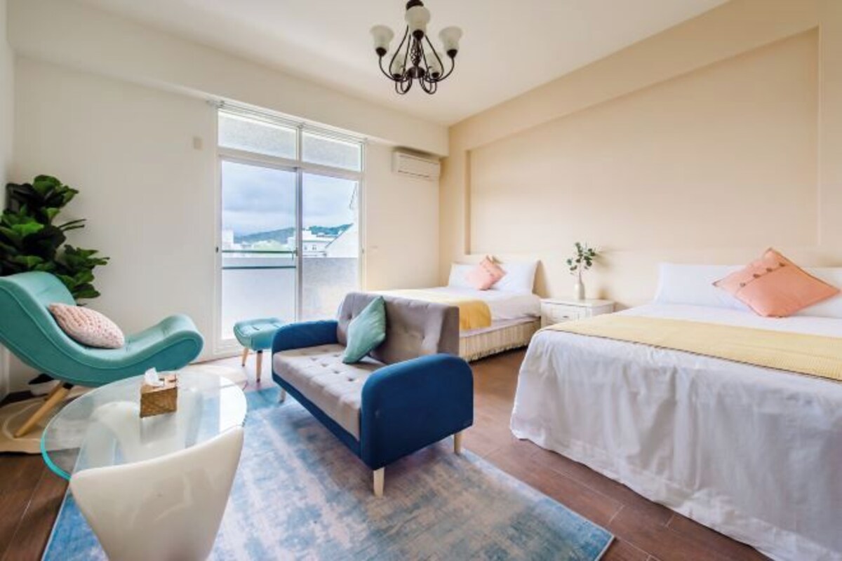A modern hotel room with a large bed, a sofa, a glass coffee table, and a view of the sea through the window.