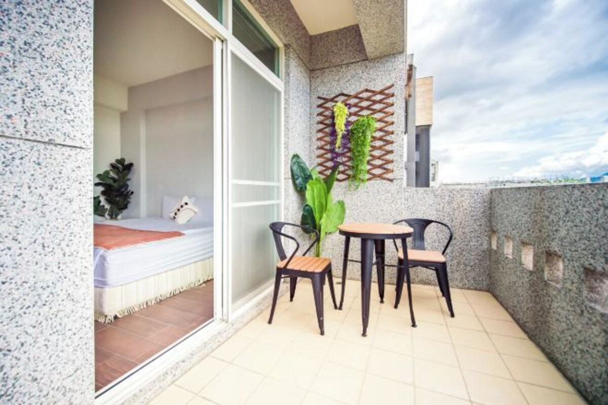 A balcony with a bed, two chairs, a table, and a potted plant.
