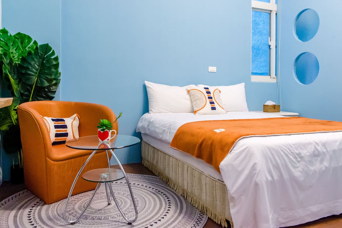 Un dormitorio con una cama, un sillón naranja, una mesita y una planta grande.