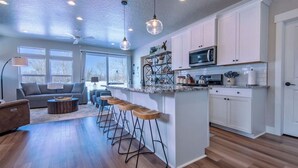 Uma cozinha moderna com armários brancos, uma ilha central com banquetas, micro-ondas e uma grande janela que proporciona iluminação natural.