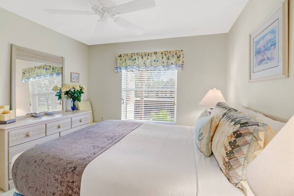 Une chambre à coucher comprenant un lit, une commode, un miroir, un ventilateur de plafond, une fenêtre avec des stores et une lampe.