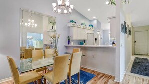 A modern dining area with a glass table, beige chairs, a chandelier, and a kitchen with decorative fish-themed items. - Active Gated Golf Community! Pool, Spa, Fitness, Tennis, Close To Shopping & Short Drive to Beaches! (Estero)