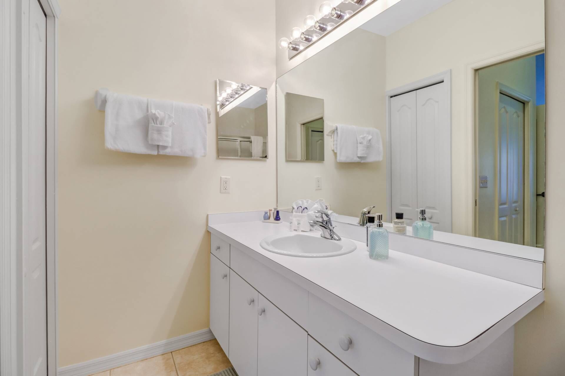 Une salle de bain moderne avec un meuble-lavabo blanc, un grand miroir et un évier. Des serviettes sont accrochées au mur et une porte est visible au fond.