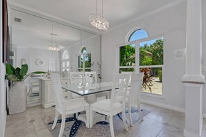 Un interior luminoso y blanco con una mesa de comedor de vidrio rodeada por sillas blancas, una araña y vistas al exterior con vegetación.