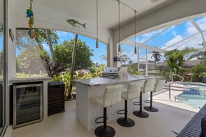 A modern outdoor kitchen with a grill, bar stools, and a poolside view. - Luxury Gulf Access Retreat | Outdoor Kitchen, Heated Pool, Hot Tub, Tiki Bar & Waterfront Dock (Cape Coral)