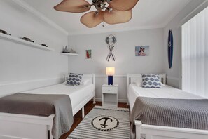 Un dormitorio con dos camas, un ventilador de techo, una mesita de noche con lámpara y una tabla de surf en la pared.