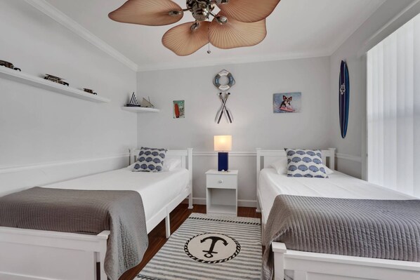 A bedroom with two beds, a ceiling fan, a nightstand with a lamp, and a surfboard on the wall.