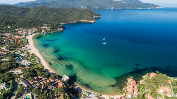 An aerial view of a coastal town with a beach, clear blue waters, and a mountain range in the background.