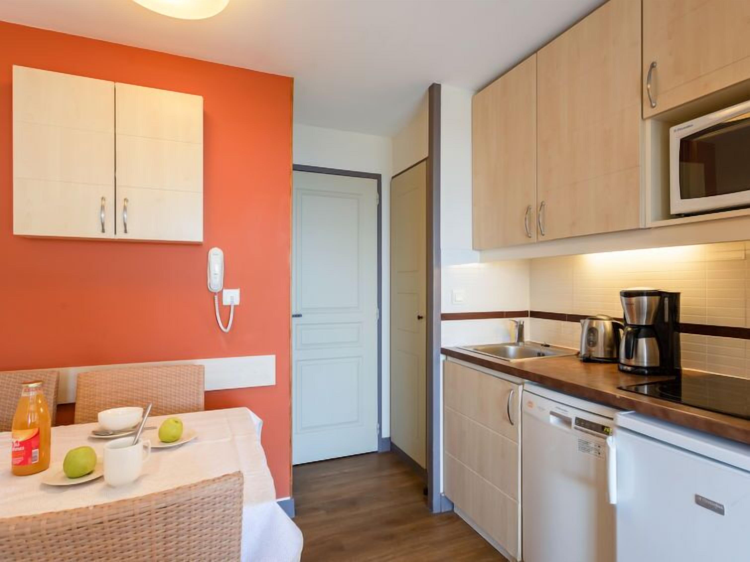 A compact kitchen with a microwave, coffee maker, and dishwasher. A dining area with a table and chairs is visible, along with a telephone on the wall.