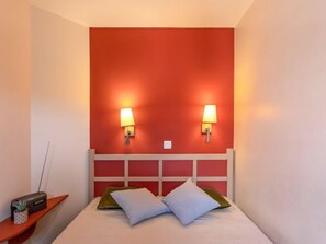 Quarto de hotel com uma parede de destaque vermelha, duas luminárias laterais, uma cabeceira branca e uma pequena mesa de cabeceira com um rádio e uma planta.