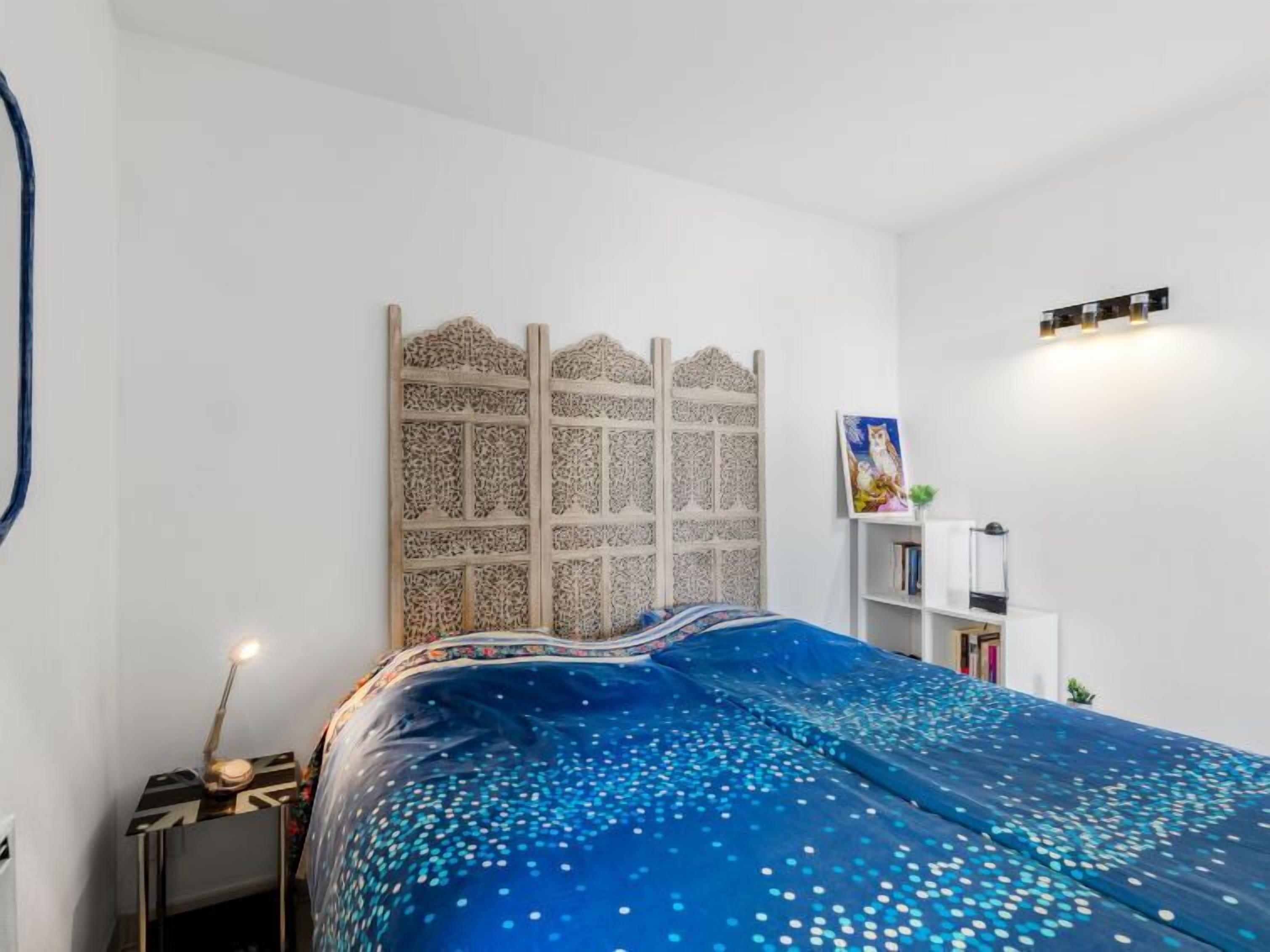 A bedroom with a bed covered in a blue blanket with white dots, a white headboard with intricate patterns, a bedside table with a lamp, and a small shelf with books and a picture frame.